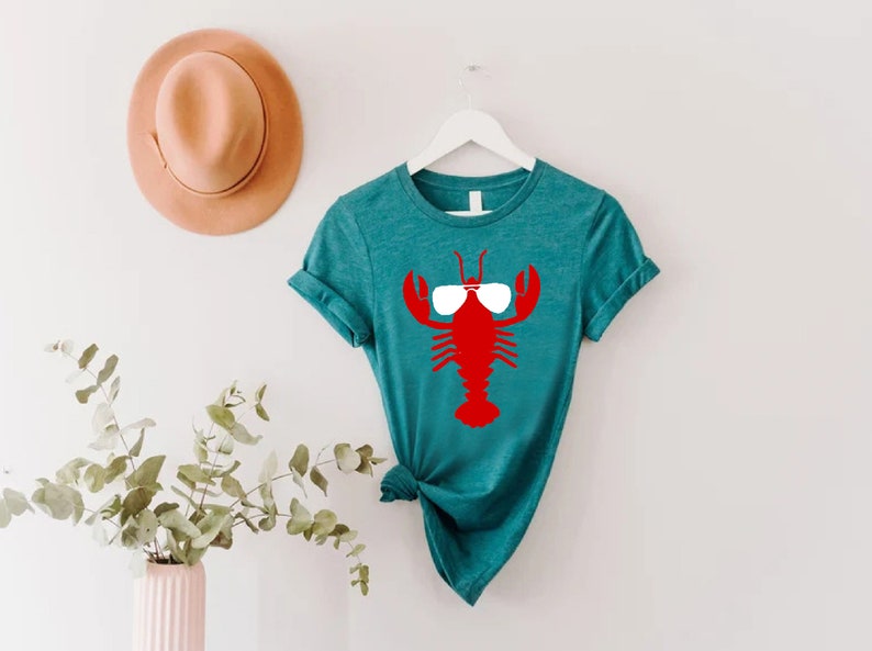 Pu&ograve; includere: T-shirt color turchese con un'aragosta rossa con occhiali da sole bianchi. La maglietta &egrave; appesa a un gancio bianco. Un cappello marrone chiaro &egrave; sulla parete a sinistra della maglietta. Un vaso con vegetazione &egrave; nell'angolo in basso a sinistra.