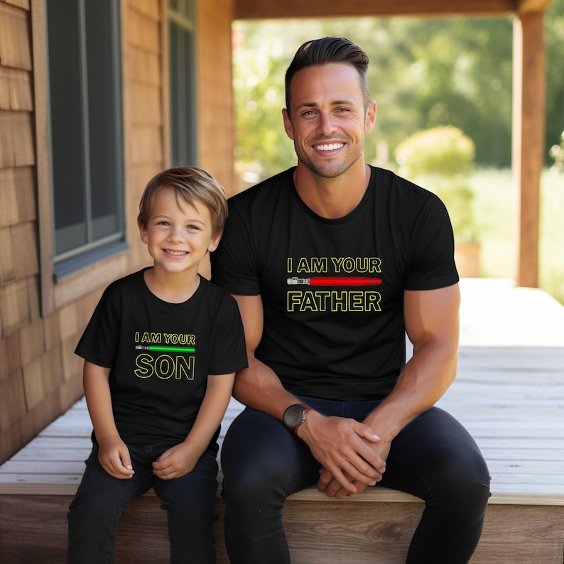 May include: A black t-shirt with the text "I am your Father" and a red lightsaber graphic. A smaller black t-shirt with the text "I am your Son" and a green lightsaber graphic. The shirts are worn by a man and a boy.