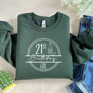 K&ouml;nnte beinhalten: Dunkelgr&uuml;nes Sweatshirt mit dem Text "21st Birthday Girl" in einem wei&szlig;en Kreisdesign. Das Design beinhaltet eine Flasche und ein Schnapsglas. Das Sweatshirt liegt neben einer Jeans.