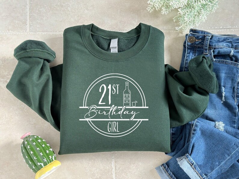 K&ouml;nnte beinhalten: Dunkelgr&uuml;nes Sweatshirt mit dem Text "21st Birthday Girl" in einem wei&szlig;en Kreisdesign. Das Design beinhaltet eine Flasche und ein Schnapsglas. Das Sweatshirt liegt neben einer Jeans.