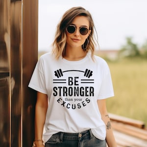 May include: White t-shirt with the text "BE STRONGER than your EXCUSES" in black. The design includes a barbell graphic above the words. The shirt is a casual style, suitable for workouts or everyday wear.