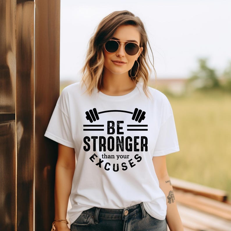 May include: White t-shirt with the text "BE STRONGER than your EXCUSES" in black. The design includes a barbell graphic above the words. The shirt is a casual style, suitable for workouts or everyday wear.