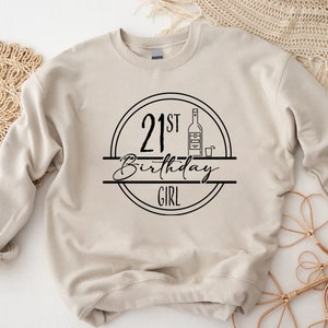 K&ouml;nnte beinhalten: Beiges Sweatshirt mit schwarzem Grafikdesign. Das Design zeigt den Text "21st Birthday Girl" in einem Kreis, mit einer Flaschen- und Schnapsglas-Illustration.