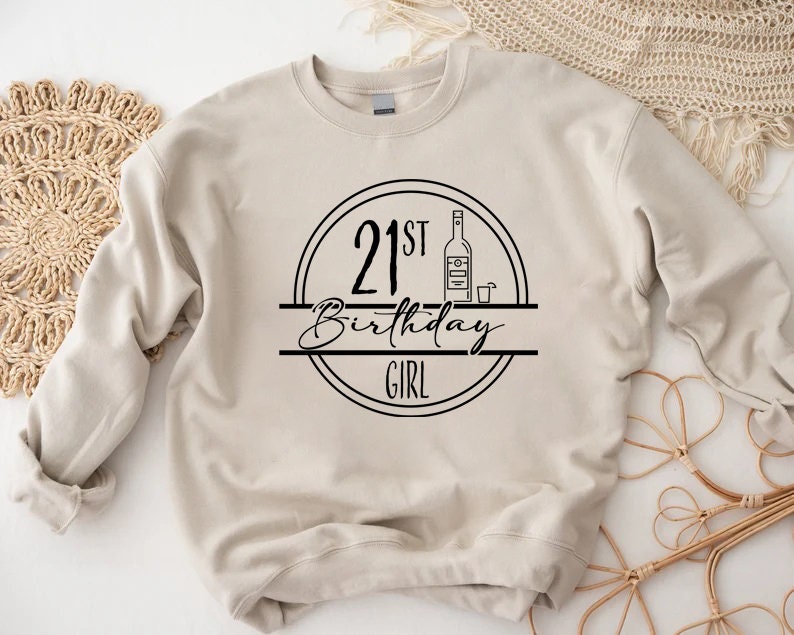 K&ouml;nnte beinhalten: Beiges Sweatshirt mit schwarzem Grafikdesign. Das Design zeigt den Text "21st Birthday Girl" in einem Kreis, mit einer Flaschen- und Schnapsglas-Illustration.