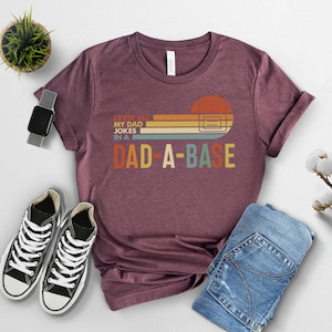 May include: Maroon t-shirt with the text "I KEEP ALL MY DAD JOKES IN A DAD-A-BASE" in a retro design. The shirt is paired with blue jeans and black and white sneakers. A smartwatch and a small potted plant are also in the image.