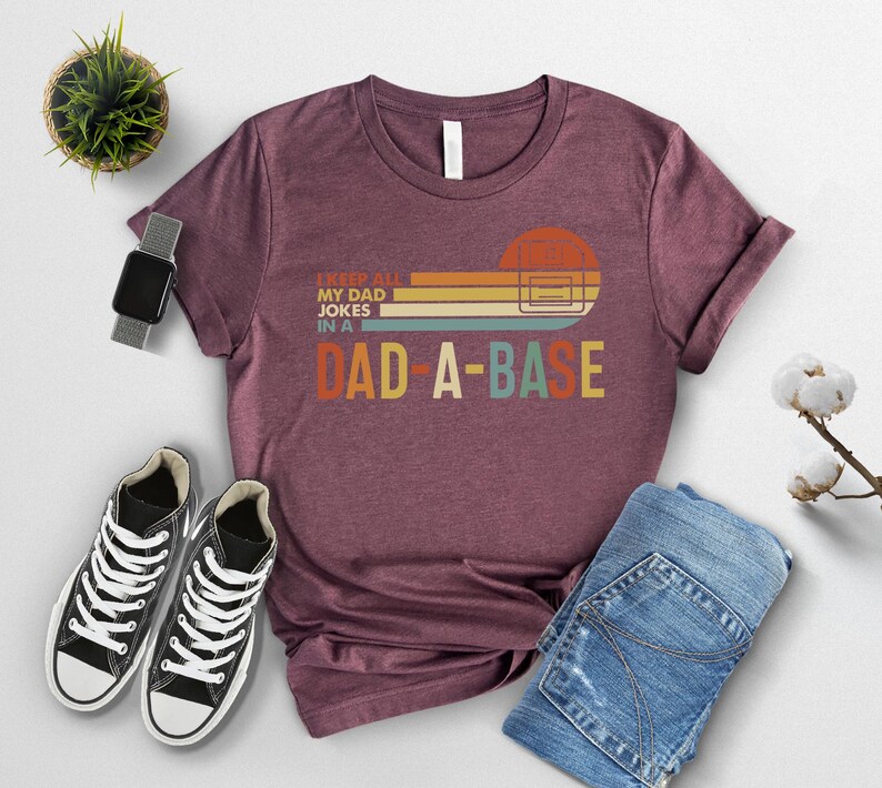 May include: Maroon t-shirt with the text "I KEEP ALL MY DAD JOKES IN A DAD-A-BASE" in a retro design. The shirt is paired with blue jeans and black and white sneakers. A smartwatch and a small potted plant are also in the image.