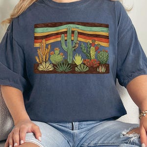 K&ouml;nnte beinhalten: Marineblaues T-Shirt mit einem W&uuml;stenmotiv. Das Design zeigt verschiedene Kakteen und einen Sonnenuntergang in Orange-, Gelb- und Gr&uuml;nt&ouml;nen. Das T-Shirt ist aus einem weichen, bequemen Material gefertigt.