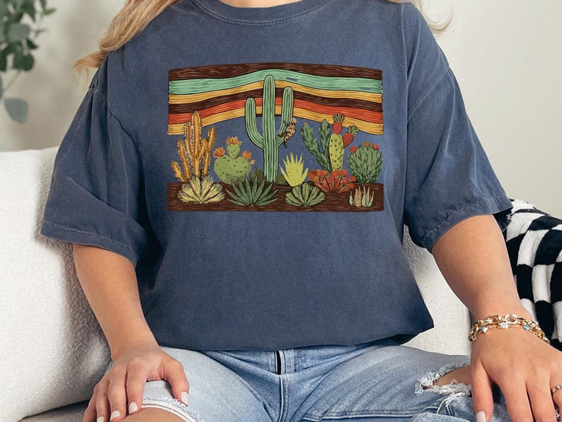 K&ouml;nnte beinhalten: Marineblaues T-Shirt mit einem W&uuml;stenmotiv. Das Design zeigt verschiedene Kakteen und einen Sonnenuntergang in Orange-, Gelb- und Gr&uuml;nt&ouml;nen. Das T-Shirt ist aus einem weichen, bequemen Material gefertigt.