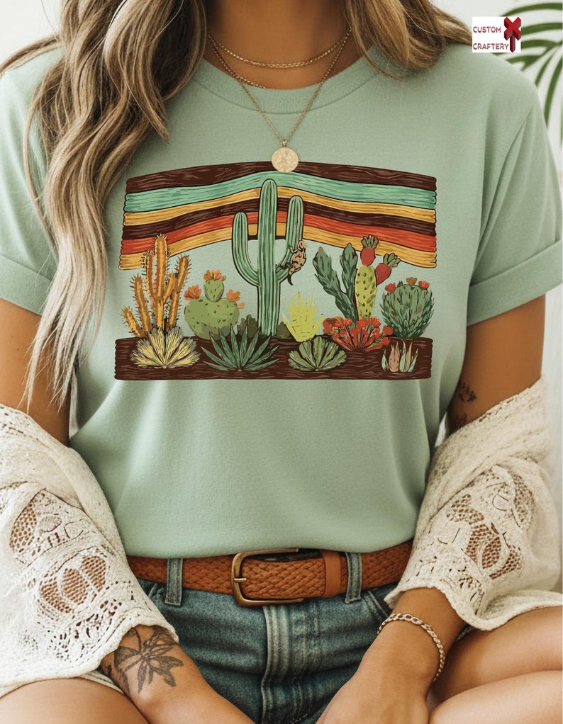 K&ouml;nnte beinhalten: Hellgr&uuml;nes T-Shirt mit einem W&uuml;stenkaktus-Design. Die Grafik zeigt verschiedene Kakteen und Sukkulenten in Gr&uuml;n, Gelb und Rot, vor einem Hintergrund aus horizontalen Streifen. Das T-Shirt hat hochgekrempelte &Auml;rmel.