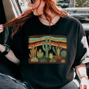 K&ouml;nnte beinhalten: Schwarzes T-Shirt mit einem W&uuml;stenkaktus-Design. Die Grafik zeigt Kakteen und Sukkulenten in Gr&uuml;n-, Gelb- und Rott&ouml;nen, vor einem Hintergrund aus horizontalen Streifen in Orange, Gr&uuml;n und Braun.
