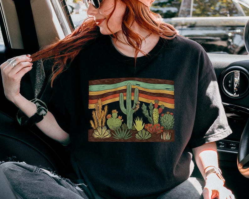 K&ouml;nnte beinhalten: Schwarzes T-Shirt mit einem W&uuml;stenkaktus-Design. Die Grafik zeigt Kakteen und Sukkulenten in Gr&uuml;n-, Gelb- und Rott&ouml;nen, vor einem Hintergrund aus horizontalen Streifen in Orange, Gr&uuml;n und Braun.