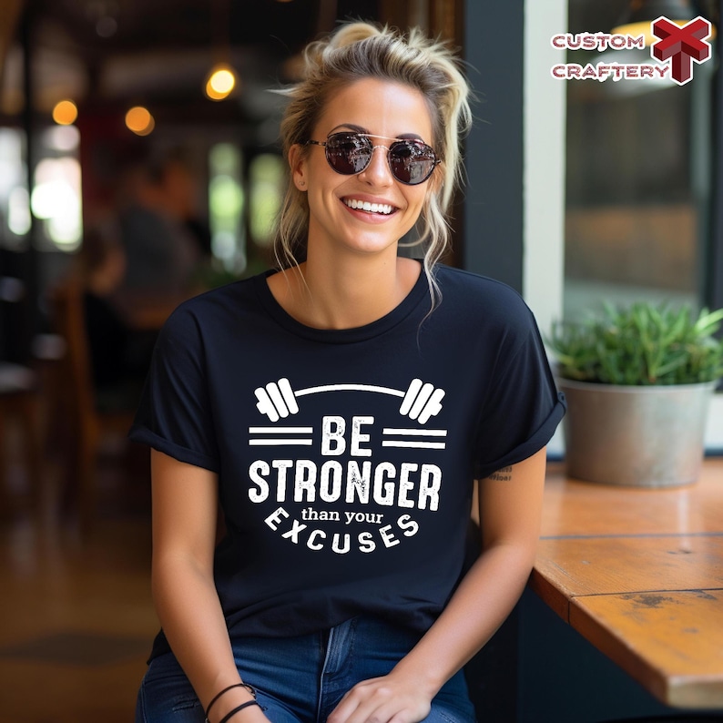 May include: Black t-shirt with the text "BE STRONGER than your EXCUSES" in white. The design includes a barbell graphic above the words. The person in the image is wearing sunglasses and blue jeans.