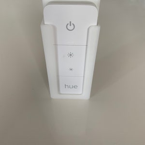 Può includere: Supporto a parete in plastica bianca per un interruttore di luce intelligente Philips Hue. L'interruttore ha un pulsante di accensione, un controllo della luminosità e la parola "hue" stampata su di esso.