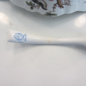 May include: A white porcelain handle with a bulbous end, possibly from a serving piece. The handle has a blue stamp with text and a decorative design. The handle is slightly curved and has a smooth, glossy finish.