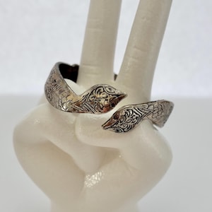 Puede incluir: Un anillo de serpiente plateado con intrincados patrones florales y geométricos. El anillo está envuelto alrededor de una mano blanca, mostrando el diseño. La cabeza y la cola de la serpiente son visibles, creando un diseño único y abierto.