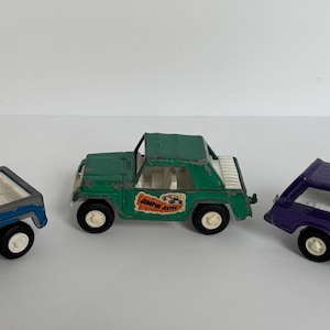 May include: Three vintage toy cars: a blue pickup truck, a green jeep-like vehicle, and a purple hatchback. Each car has white wheels and colorful decals. The blue truck shows signs of wear.