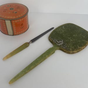 May include: An orange and white round tin box with a floral design, a green-handled letter opener, and a green hand mirror with a decorative design. The items are arranged on a white surface.