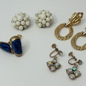 May include: Assortment of vintage earrings, including a pair of blue enamel clip-on earrings, a pair of gold hoop earrings with a textured design, and two pairs of clip-on earrings with white and iridescent stones.