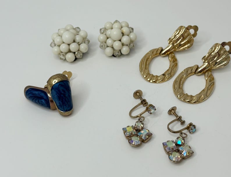 May include: Assortment of vintage earrings, including a pair of blue enamel clip-on earrings, a pair of gold hoop earrings with a textured design, and two pairs of clip-on earrings with white and iridescent stones.