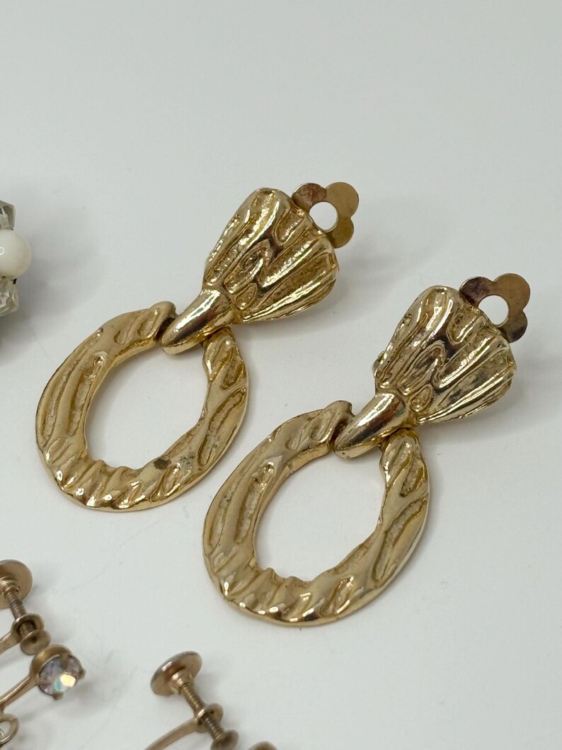 May include: A pair of gold tone clip-on earrings with a textured oval design and a decorative top.
