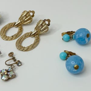 May include: A set of vintage earrings featuring a pair of gold tone clip-on earrings with a textured oval design, a pair of blue and white clip-on earrings with a round design, and a pair of silver tone clip-on earrings with a crystal design.