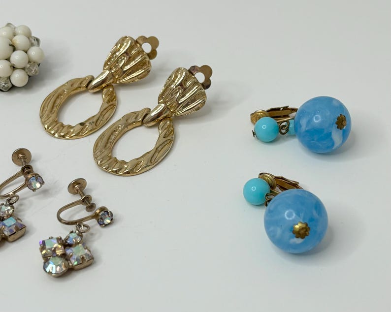 May include: A set of vintage earrings featuring a pair of gold tone clip-on earrings with a textured oval design, a pair of blue and white clip-on earrings with a round design, and a pair of silver tone clip-on earrings with a crystal design.