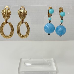May include: Two pairs of earrings. The first pair is gold with a textured oval design. The second pair is silver with a blue and turquoise bead design.