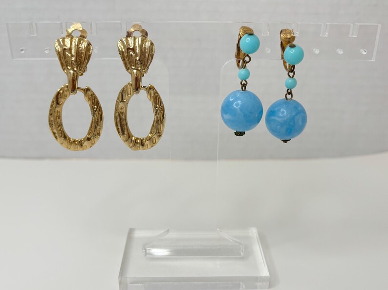 May include: Two pairs of earrings. The first pair is gold with a textured oval design. The second pair is silver with a blue and turquoise bead design.