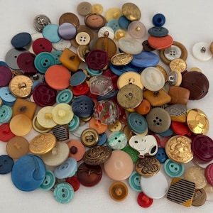 May include: A collection of assorted buttons in various colors, shapes, and sizes. The buttons include round, square, and novelty designs, with colors ranging from red, blue, and green to gold, brown, and cream. Some buttons have decorative patterns.