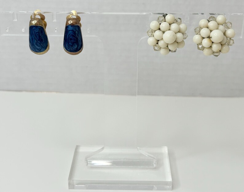 May include: Two pairs of earrings on a display stand. One pair is blue with a swirl pattern and the other pair is white and round.