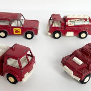 May include: Four vintage toy vehicles in shades of red, including a truck, a tanker, a jeep, and a military-style vehicle. The toys have white and yellow accents and black wheels. The jeep has a yellow sticker.