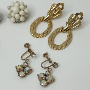 May include: Three pairs of vintage earrings. The first pair is gold with a textured oval design. The second pair is gold with a cluster of iridescent rhinestones. The third pair is white with a cluster of small white beads.