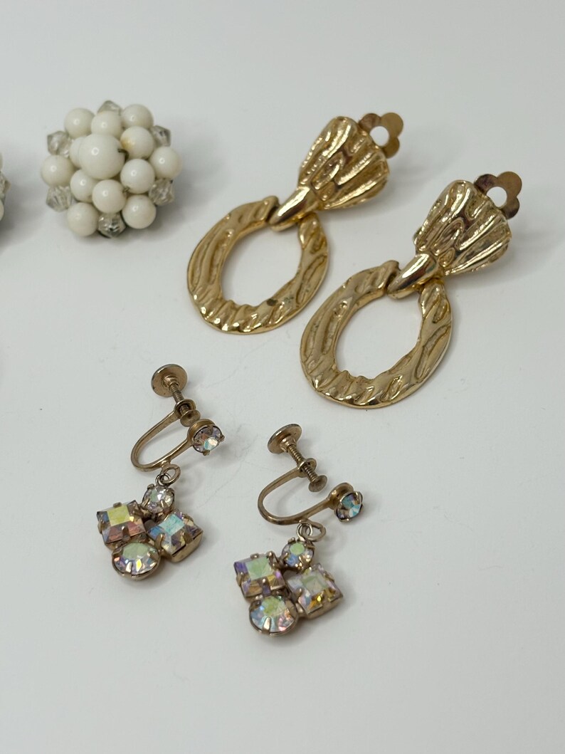 May include: Three pairs of vintage earrings. The first pair is gold with a textured oval design. The second pair is gold with a cluster of iridescent rhinestones. The third pair is white with a cluster of small white beads.