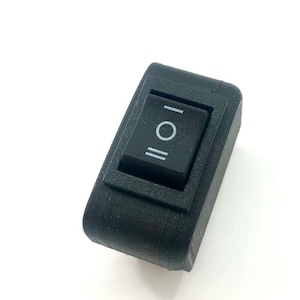 May include: A black rectangular rocker switch with a rectangular button. The button has a white circle between two parallel white lines, indicating the on/off positions. The switch is set against a white background.