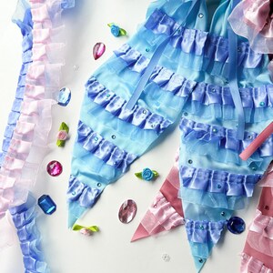 K&ouml;nnte beinhalten: Zwei blaue und rosa Stoffschleifen mit gekr&auml;uselten R&auml;ndern und Strasssteinen. Die Schleifen befinden sich auf einem wei&szlig;en Hintergrund mit verstreuten Strasssteinen und kleinen Blumen.