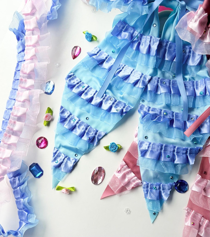 K&ouml;nnte beinhalten: Zwei blaue und rosa Stoffschleifen mit gekr&auml;uselten R&auml;ndern und Strasssteinen. Die Schleifen befinden sich auf einem wei&szlig;en Hintergrund mit verstreuten Strasssteinen und kleinen Blumen.