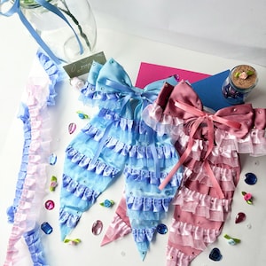 K&ouml;nnte beinhalten: Zwei gro&szlig;e, dekorative Schleifen mit langen B&auml;ndern. Die Schleifen bestehen aus mehreren Lagen T&uuml;ll und Satinband. Eine Schleife ist hellblau und die andere rosa. Beide Schleifen sind mit Strasssteinen verziert.