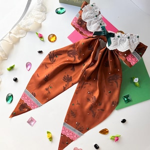 May include: An orange hair bow with a floral pattern and embellished with small, colorful rhinestones. The bow features a white lace trim and a green ribbon. The ends are trimmed with pink lace and black and white checkered fabric.