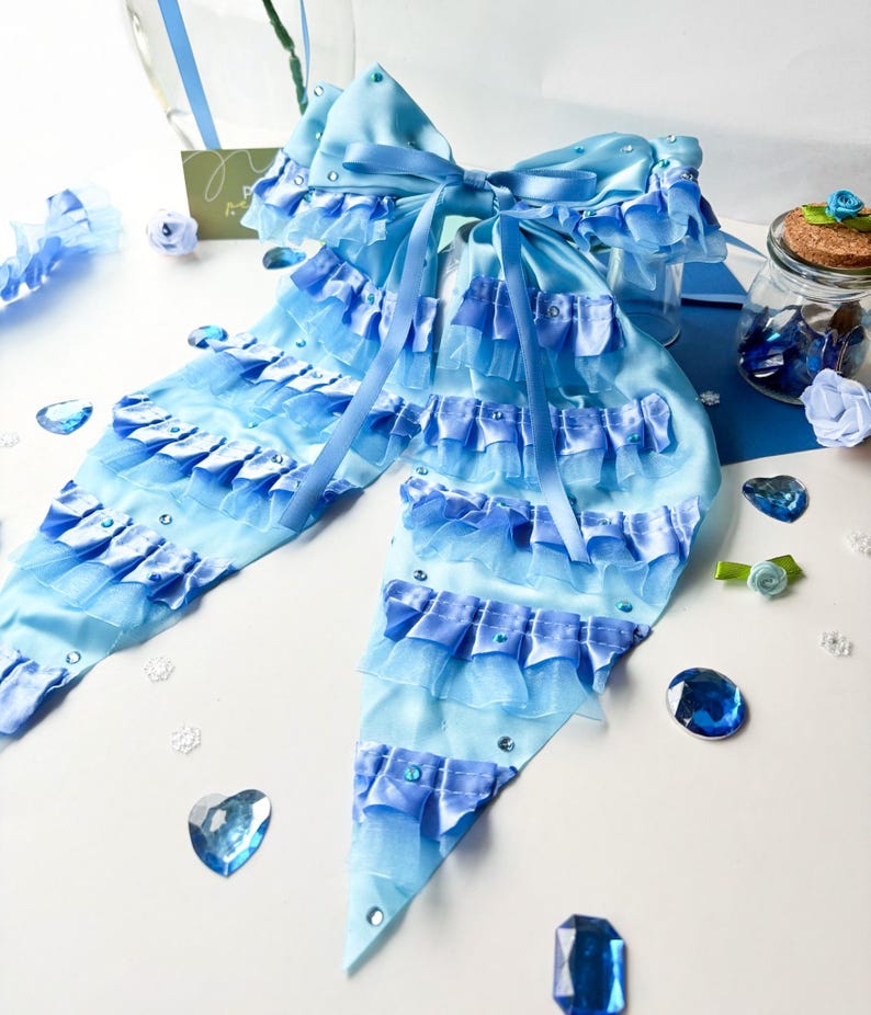 May include: A large blue and light blue bow with multiple layers of ruffles and rhinestones. The bow is tied in a large knot and has long tails.