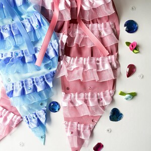 K&ouml;nnte beinhalten: Zwei Stoff-Meerjungfrauen-Schw&auml;nze, einer blau und einer rosa, mit mehreren R&uuml;schen und Strasssteinen. Die Schw&auml;nze liegen auf einer wei&szlig;en Oberfl&auml;che mit verschiedenen blauen und rosa Edelsteinen.