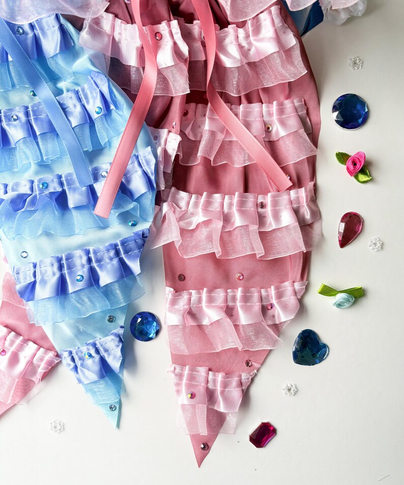 K&ouml;nnte beinhalten: Zwei Stoff-Meerjungfrauen-Schw&auml;nze, einer blau und einer rosa, mit mehreren R&uuml;schen und Strasssteinen. Die Schw&auml;nze liegen auf einer wei&szlig;en Oberfl&auml;che mit verschiedenen blauen und rosa Edelsteinen.