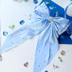 May include: A light blue satin bow with silver glitter and rhinestones. The bow is tied in a large loop with two long tails. The bow is on a white surface with other decorative items.