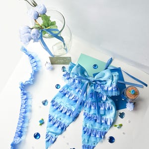 May include: A light blue satin bow with multiple layers of ruffles and rhinestones. The bow is tied with a light blue ribbon and is surrounded by blue and clear gemstones.