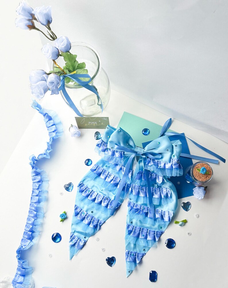 May include: A light blue satin bow with multiple layers of ruffles and rhinestones. The bow is tied with a light blue ribbon and is surrounded by blue and clear gemstones.