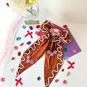 May include: A brown hair bow with a gingerbread cookie design, featuring white icing details and candy accents. The bow is decorated with red and white swirl candies and small, colorful gems. The overall theme is festive and whimsical.