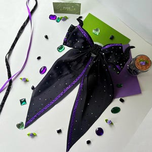 May include: A black satin bow with purple trim and scattered rhinestones. The bow has long, pointed tails and is surrounded by decorative purple and green gems, ribbons, and small floral accents. The bow is likely a hair accessory.