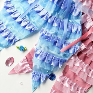 K&ouml;nnte beinhalten: Zwei Stoffhaarb&auml;nder, eines hellblau und eines rosa, mit gekr&auml;uselten R&auml;ndern und Strasssteinen. Die Schleifen bestehen aus mehreren Lagen T&uuml;ll und Satinband.