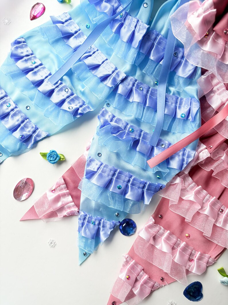 K&ouml;nnte beinhalten: Zwei Stoffhaarb&auml;nder, eines hellblau und eines rosa, mit gekr&auml;uselten R&auml;ndern und Strasssteinen. Die Schleifen bestehen aus mehreren Lagen T&uuml;ll und Satinband.