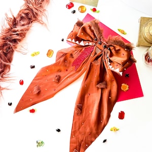 May include: A large, brown hair bow with a decorative design. The bow features a textured, fur-like trim and is embellished with small, sparkling accents. Scattered around the bow are colorful, gem-like decorations and a small card with gold text.