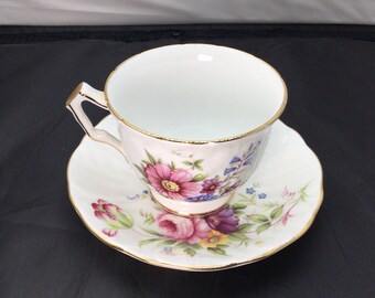 May include: A white teacup and saucer with a floral pattern. The cup has a gold rim and the saucer has a gold rim and a scalloped edge. The flowers are pink, purple, and blue.